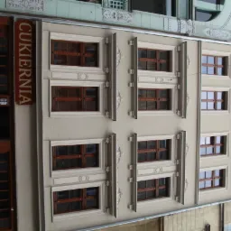 Fasada kamienicy z oknami w ozdobnych ramach, widoczny szyld 'Cukiernia' z boku. Architektura miejska, jasna kolorystyka elewacji, detale dekoracyjne.