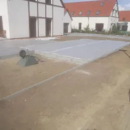 Przygotowanie podłoża pod nawierzchnię na posesji: wyrównany teren, obrzeża z kostki, rozłożona geowłóknina, taczka z narzędziami w tle domy jednorodzinne.