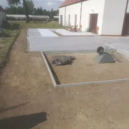 Przygotowanie terenu pod budowę tarasu: wyrównany teren z obrzeżami, przykryty folią, taczka i worki z materiałem budowlanym w tle, widok na dom z tarasem i meblami ogrodowymi.