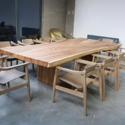 Stół z blatem z litego drewna o nieregularnych krawędziach, otoczony drewnianymi krzesłami w minimalistycznym stylu, na tle betonowej kolumny i szklanej ściany.