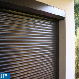 Zewnętrzna roleta zwinięta, z widocznym mechanizmem w kolorze ciemnego brązu, zamontowana na jasnej elewacji budynku z zielenią w tle.