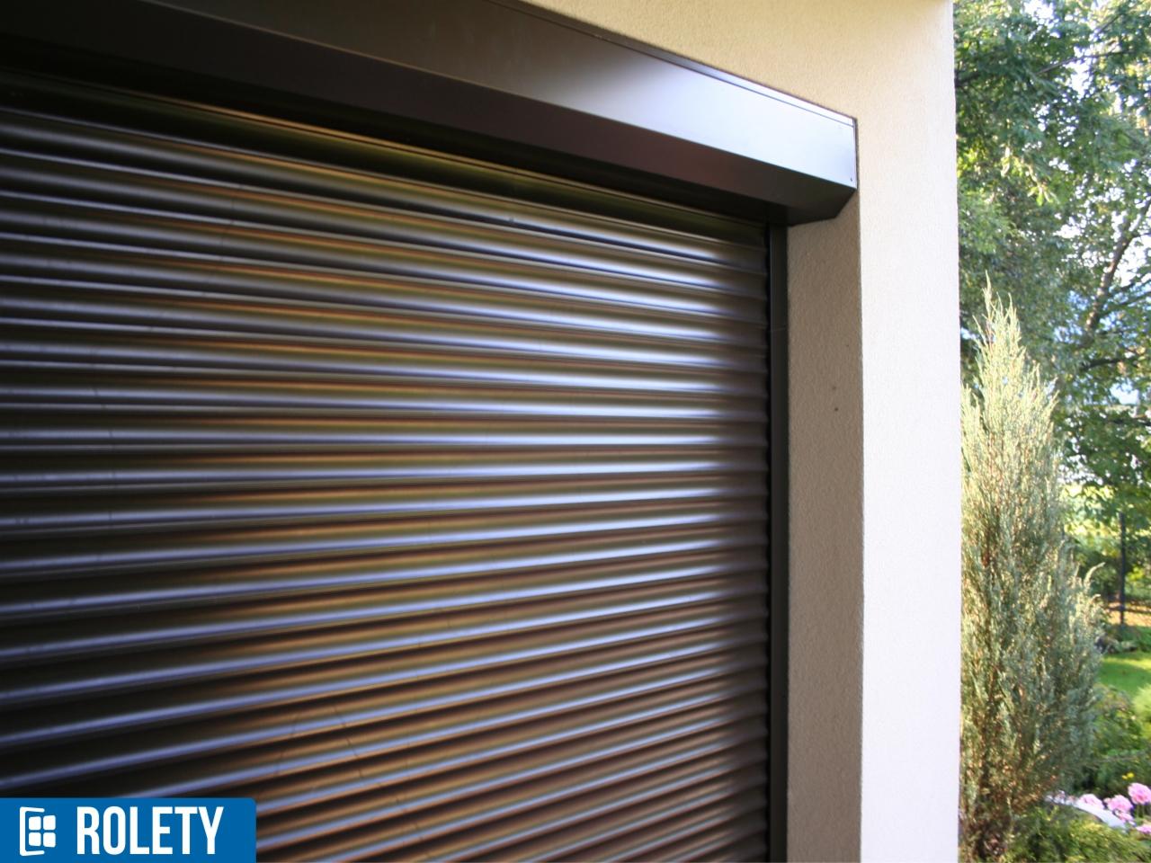 Zewnętrzna roleta zwinięta, z widocznym mechanizmem w kolorze ciemnego brązu, zamontowana na jasnej elewacji budynku z zielenią w tle.