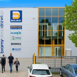 Budynek firmy Bilbo z ekspozycją okien i bram. Na fasadzie logotypy producentów. Rodzina przechodzi obok szlabanu i zaparkowanych aut, podnosząc atrakcyjność wizualną oferty.
