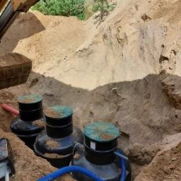 Montaż trzech czarnych zbiorników z zielonymi pokrywami w wykopie ziemnym. Widoczny fragment koparki i niebieski wąż. Instalacja w trakcie realizacji.