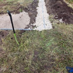 Darmont - Drenaż liniowy: rów z ułożoną rurą drenażową zasypany białym żwirem, wyrównana ziemia po bokach, w tle trawa, sprzęt budowlany i osoba. Widok z góry.