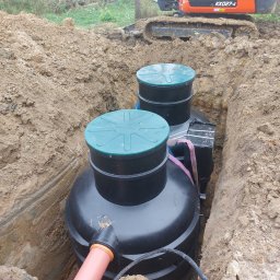 Darmont - Montaż dwukomorowej, czarnej oczyszczalni z zielonymi pokrywami w wykopie. Pomarańczowa rura i koparka Kubota w tle. Etap instalacji.