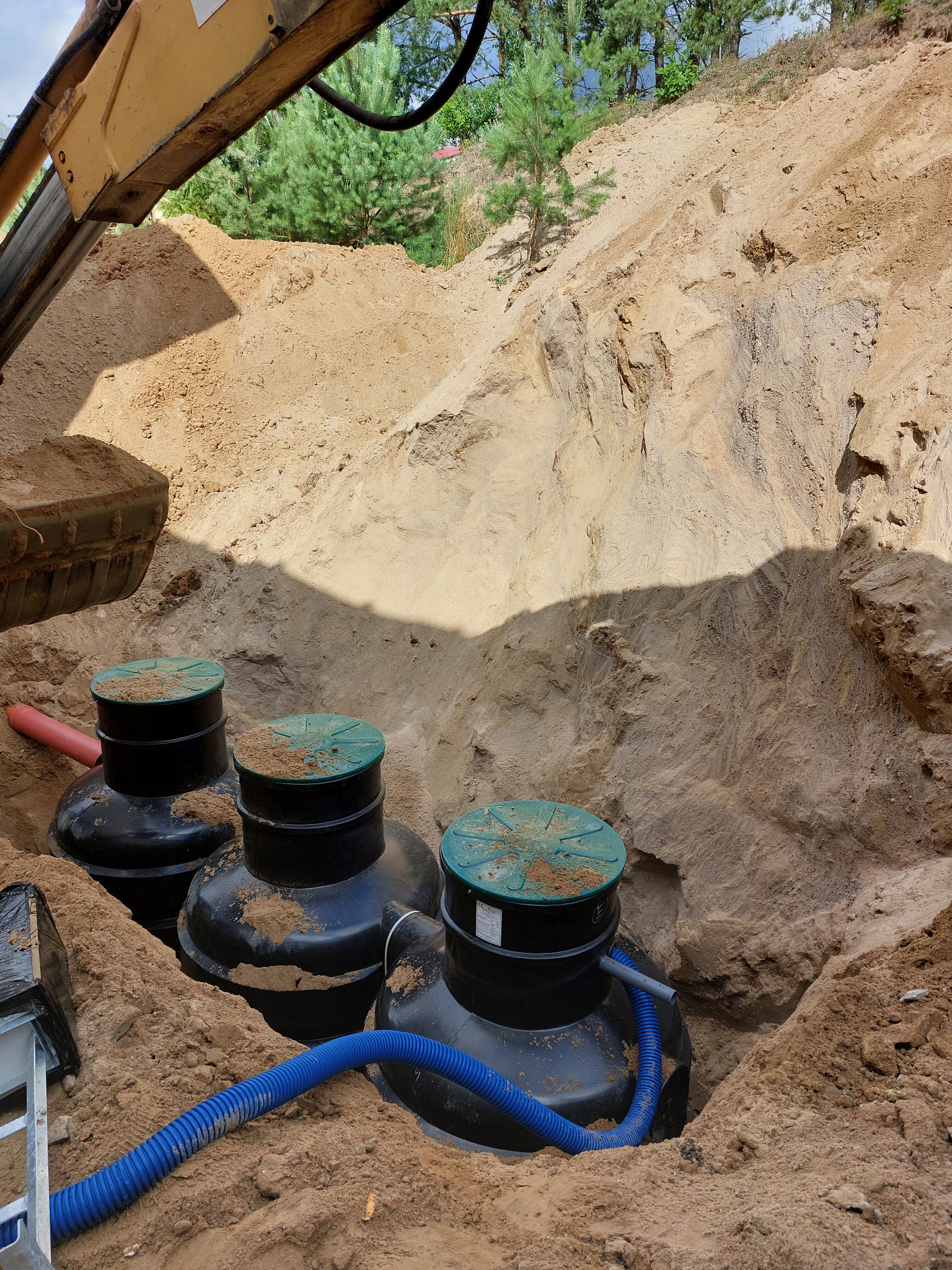 Montaż trzech czarnych zbiorników z zielonymi pokrywami w wykopie ziemnym. Widoczny fragment koparki i niebieski wąż. Instalacja w trakcie realizacji.