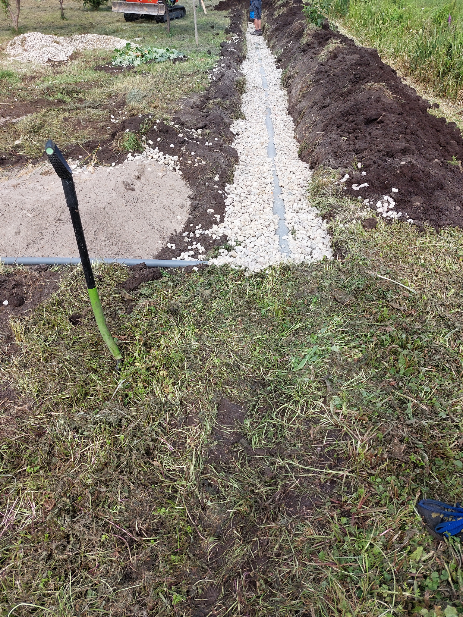 Drenaż liniowy: rów z ułożoną rurą drenażową zasypany białym żwirem, wyrównana ziemia po bokach, w tle trawa, sprzęt budowlany i osoba. Widok z góry.