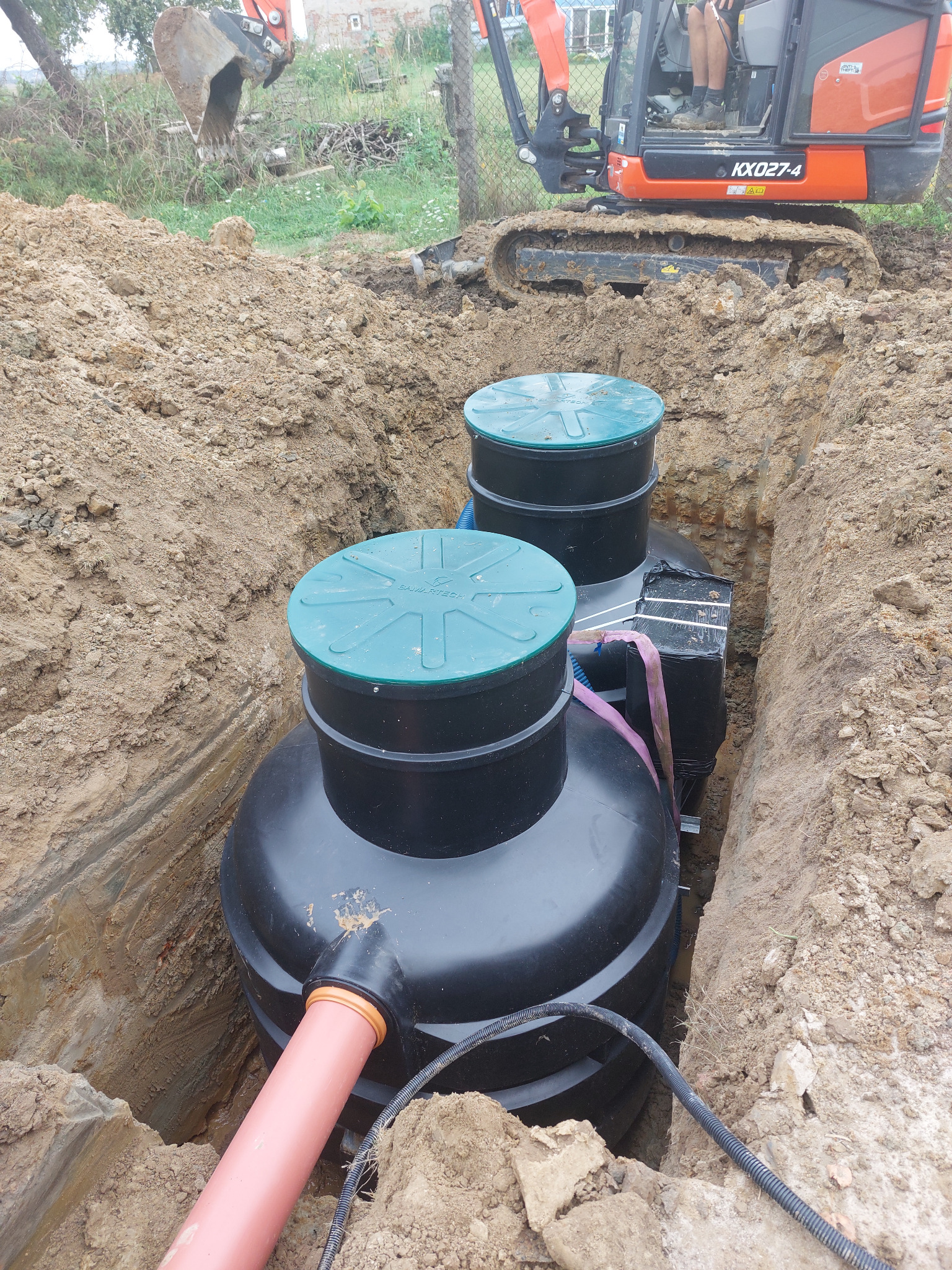 Montaż dwukomorowej, czarnej oczyszczalni z zielonymi pokrywami w wykopie. Pomarańczowa rura i koparka Kubota w tle. Etap instalacji.