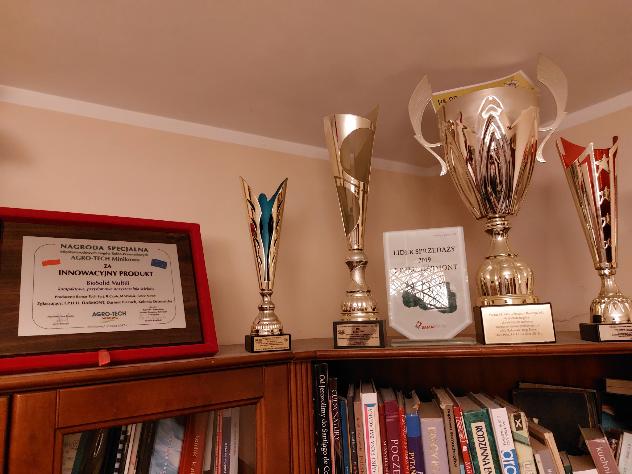 Półka z nagrodami i certyfikatem 'Innowacyjny Produkt' dla BioSolid Multi8, kompaktowej oczyszczalni ścieków. Widoczne książki i puchary, w tle jasna ściana.