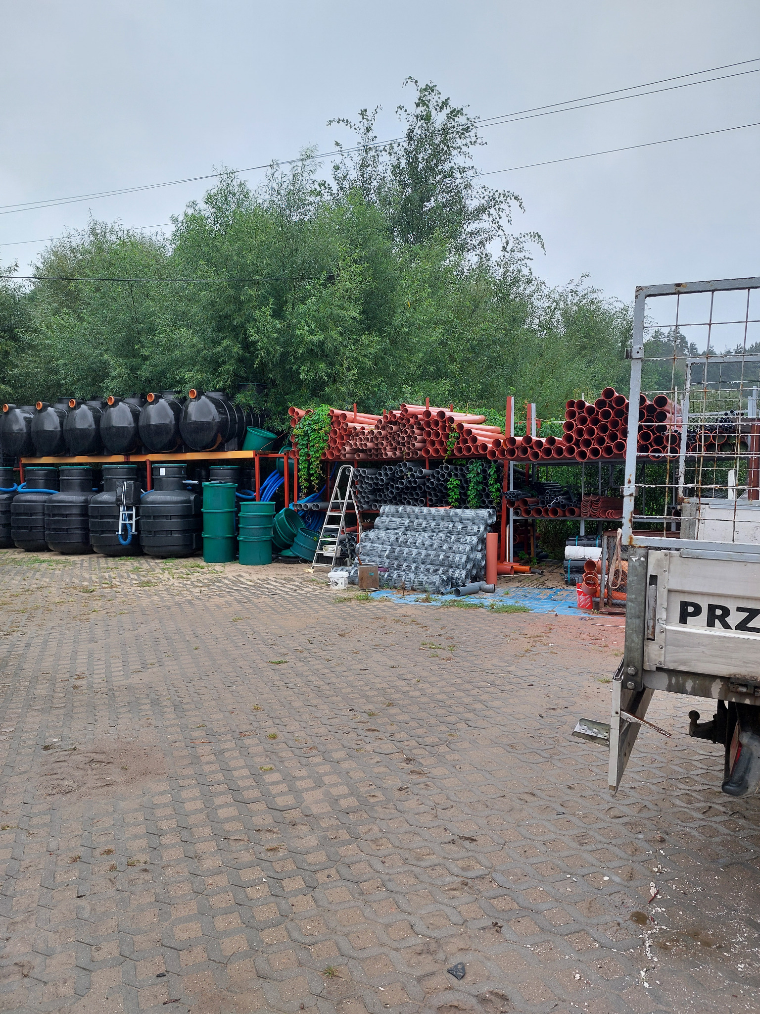 Magazyn rur i zbiorników z tworzyw sztucznych na zewnątrz, widok z boku przyczepy. Różne kolory i rozmiary rur ułożone na regałach.