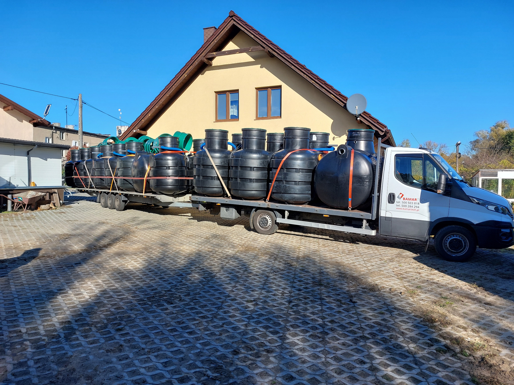 Transport czarnych zbiorników na przyczepie ciężarówki Bamartech na tle domu z brązowym dachem i błękitnego nieba. Zbiorniki zabezpieczone pomarańczowymi pasami.