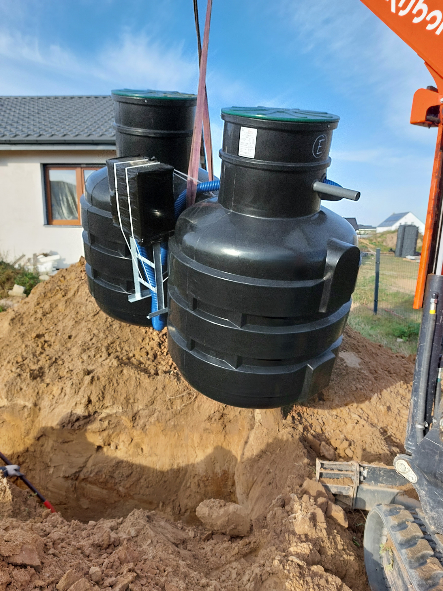 Montaż dwukomorowej, czarnej oczyszczalni ścieków w wykopie za pomocą dźwigu. Widoczny fragment domu i niebo.