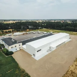 Widok z lotu ptaka na kompleks budynków: starszy budynek biurowy z niebieską elewacją i nowa hala magazynowa z białym dachem, otoczone parkingiem i terenem zielonym.