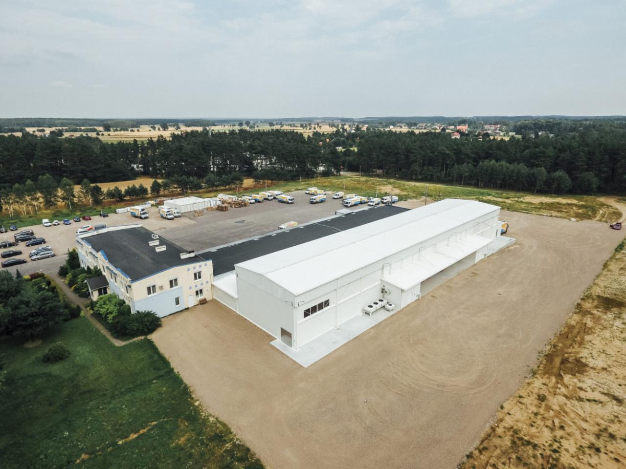 Widok z lotu ptaka na kompleks budynków: starszy budynek biurowy z niebieską elewacją i nowa hala magazynowa z białym dachem, otoczone parkingiem i terenem zielonym.