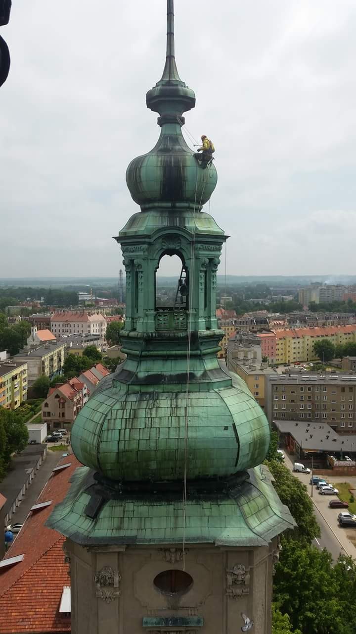 Pracownik zabezpieczony linami, wykonuje prace na szczycie wieży kościoła z zieloną kopułą, widok na miasto w tle.