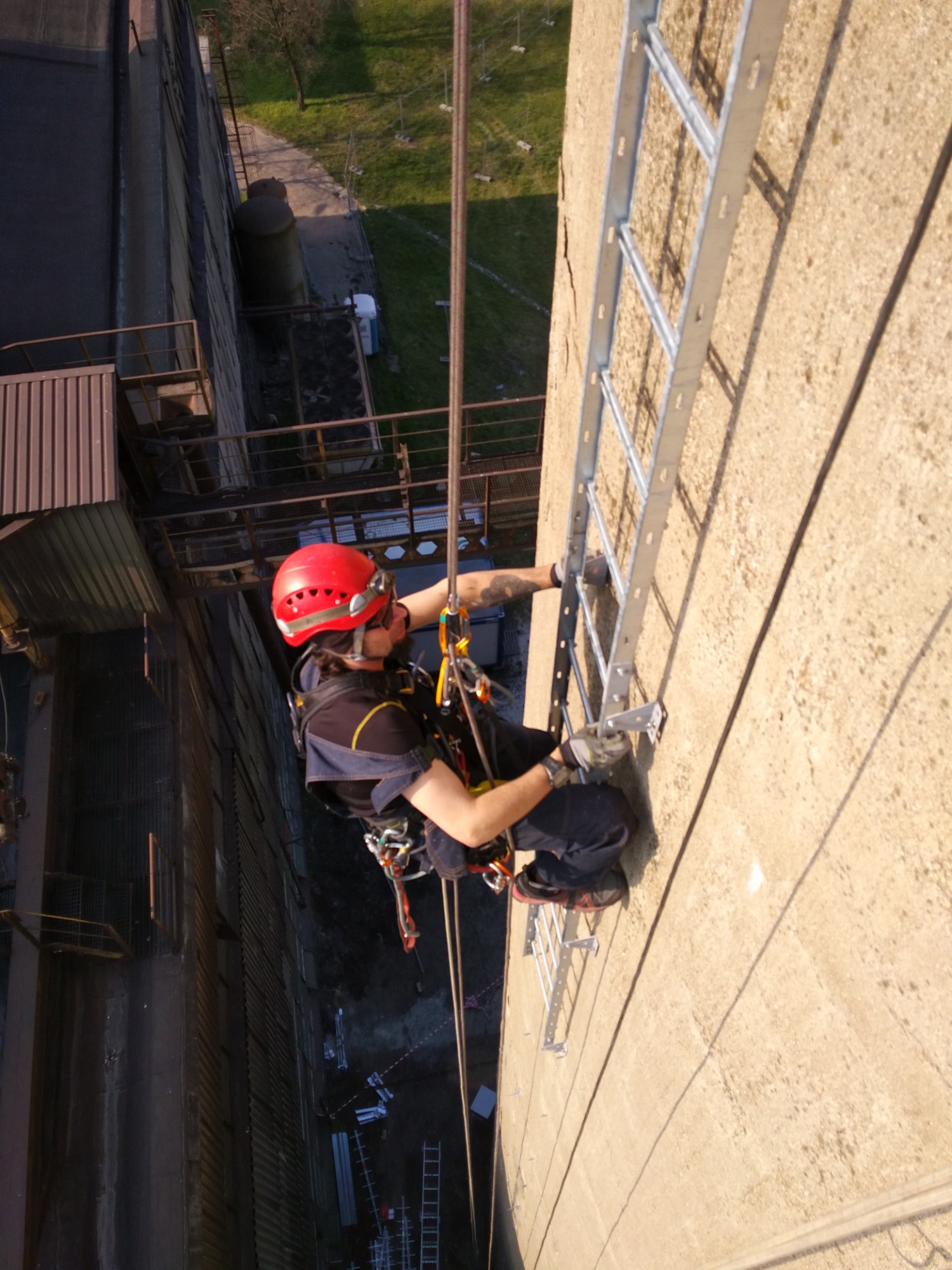 Monter w czerwonym kasku, zabezpieczony linami, instaluje drabinkę na bocznej ścianie wysokiego komina przemysłowego. Widok z góry.