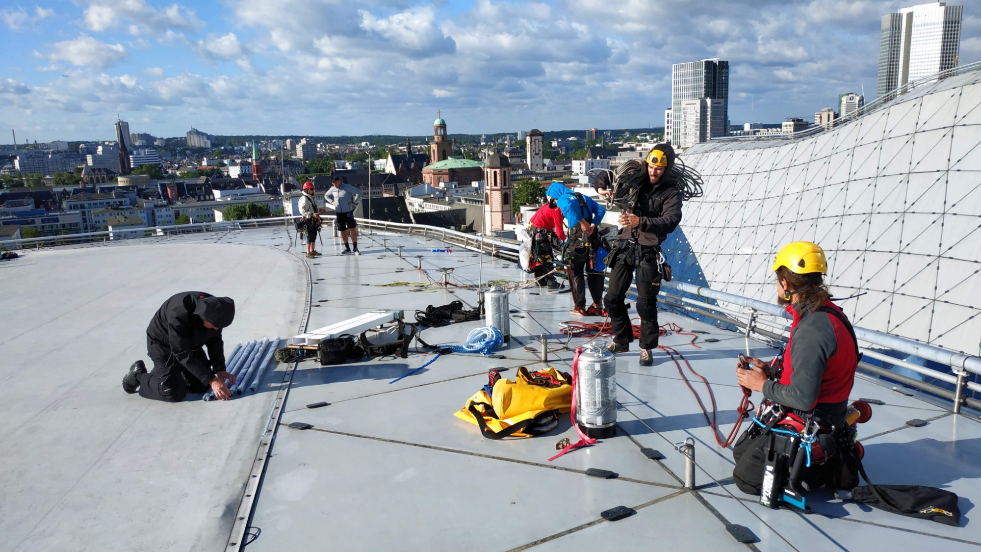 Grupa alpinistów przemysłowych na dachu budynku w trakcie pracy, widok na miasto w tle.