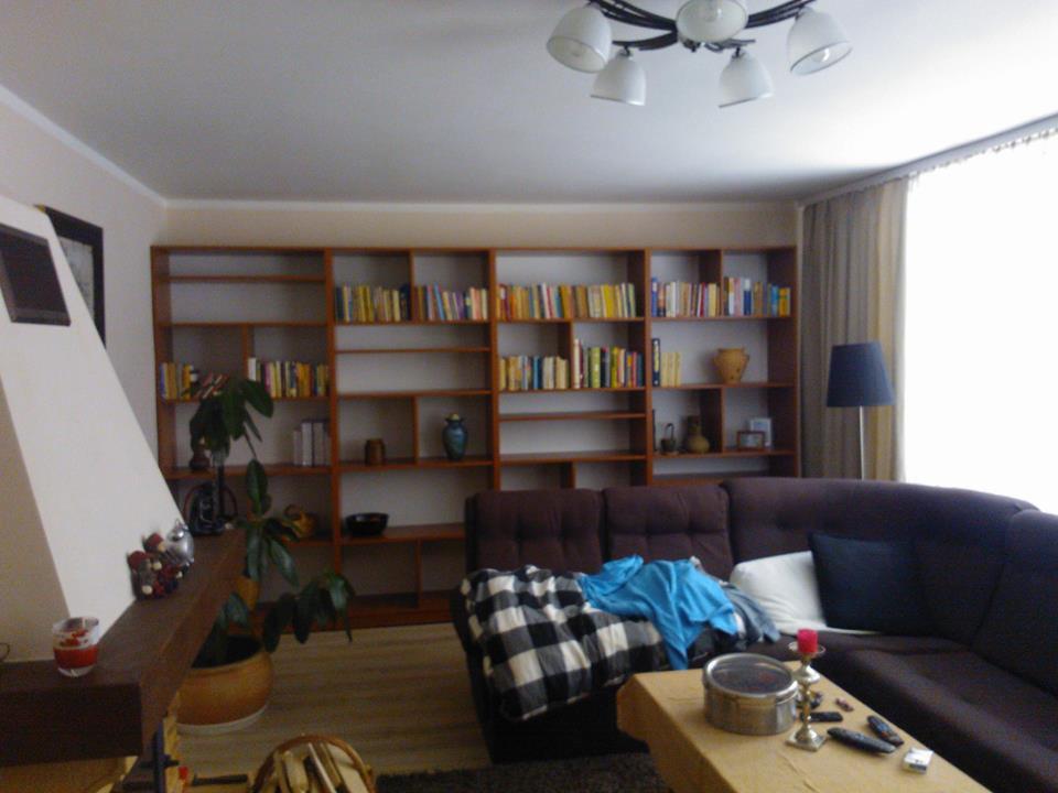 Przestronny salon z dużą, drewnianą biblioteczką wypełnioną książkami. W tle okno z jasnymi zasłonami i ciemna sofa z poduszkami. Na pierwszym planie stolik kawowy.