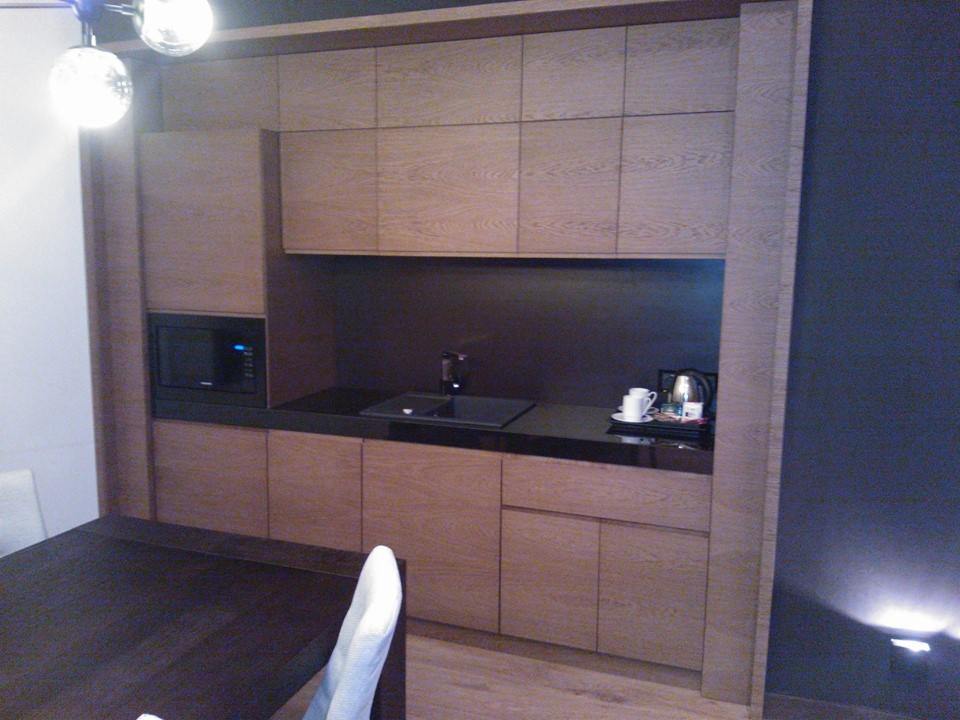 Drewniana zabudowa kuchenna z mikrofalówką i czarnym blatem, uzupełniona o czajnik i filiżanki, widoczna z perspektywy stołu. Nowoczesny design.