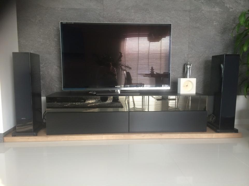 Nowoczesny salon z czarną szafką RTV pod telewizorem, głośnikami po bokach i szarą ścianą w tle. Minimalistyczny design z elementami dekoracyjnymi.