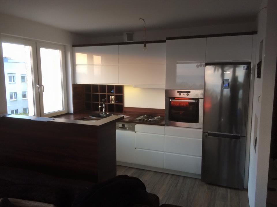 Nowoczesna, jasna kuchnia z białymi szafkami i drewnianym blatem. Widoczny zlew, płyta grzewcza, piekarnik i lodówka. Na blacie przy oknie leżą dokumenty.