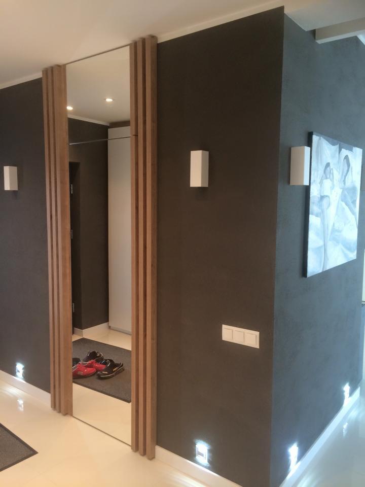 Nowoczesny przedpokój z dużym lustrem obramowanym drewnianymi listwami, oświetlony punktowymi lampami podłogowymi i ściennymi. Na ścianie obraz w stonowanych barwach.