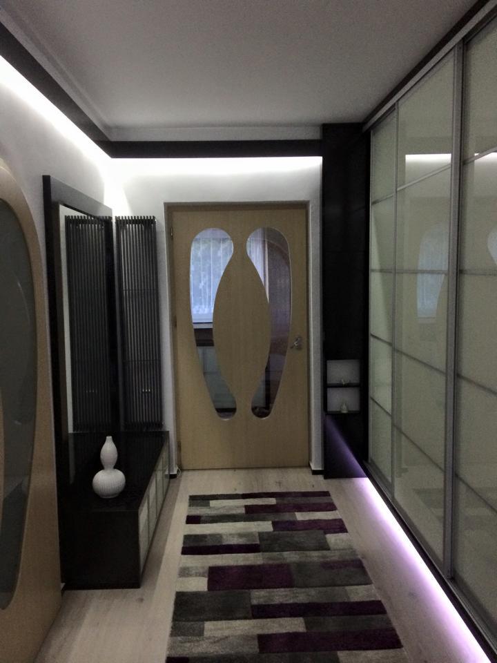 Nowoczesny przedpokój z podświetlaną szafą wnękową, designerskimi drzwiami z przeszkleniem i dywanem w geometryczne wzory. Minimalistyczna konsola z wazonem.