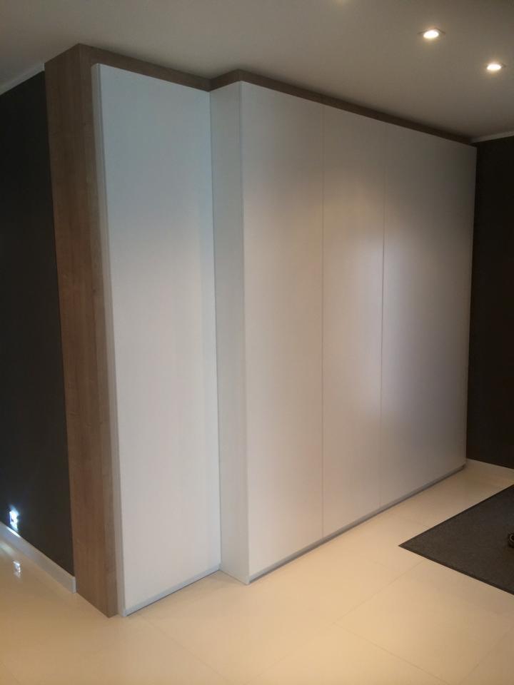 Minimalistyczna szafa wnękowa z białymi frontami, wykończona drewnianą ramą, w nowoczesnym wnętrzu z subtelnym oświetleniem punktowym i ciemną ścianą.