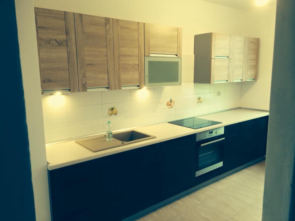 Nowoczesna kuchnia z drewnianymi szafkami górnymi i ciemnymi dolnymi, biały blat, płyta indukcyjna i piekarnik. Na ścianie białe płytki z delikatnym wzorem.