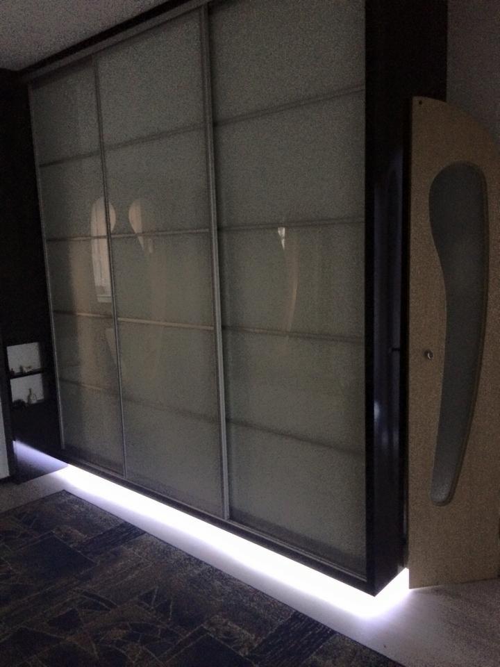 Nowoczesna szafa wnękowa z przesuwnymi, mlecznymi drzwiami i subtelnym oświetleniem LED u dołu, tworzącym nastrojowy klimat w ciemnym pomieszczeniu.