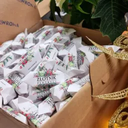Pudełko wypełnione kr&oacute;wkami reklamowymi 'Złotnik Żeromscy' z motywem świątecznym, widoczne logo 'Wrona' na pudełku, dekoracja świąteczna w tle.
