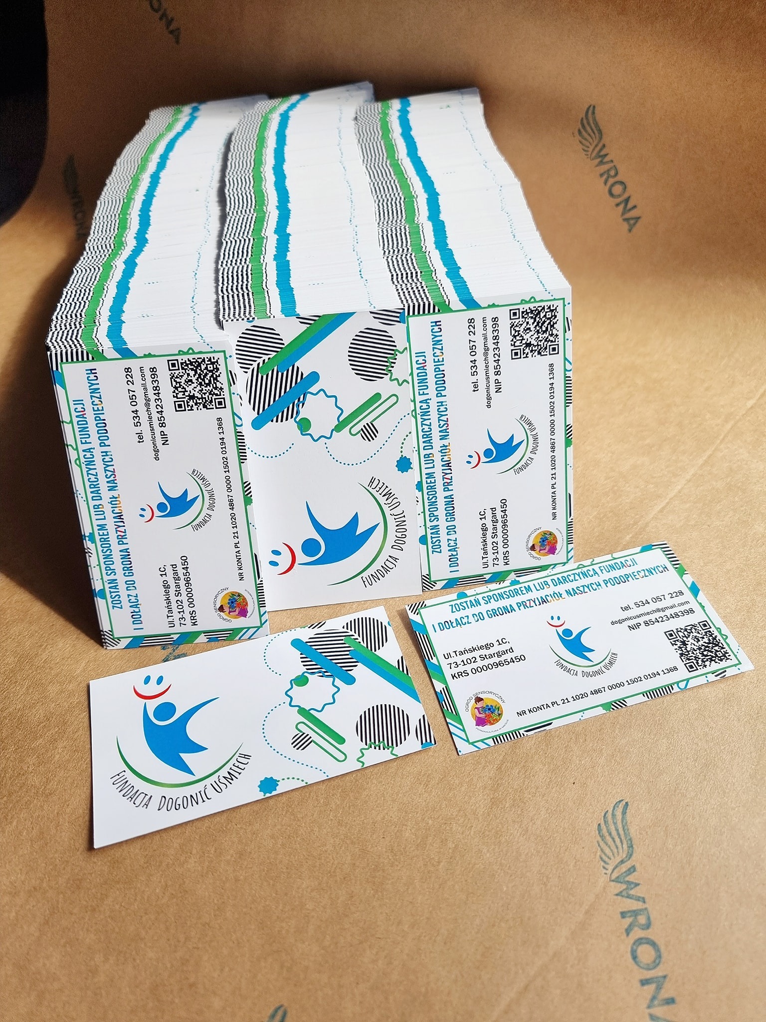 Stos wizytówek Fundacji Dogonić Uśmiech z niebiesko-zielonym logo i danymi kontaktowymi, leżące na tekturowym tle z widocznym fragmentem logotypu drukarni Wrona.