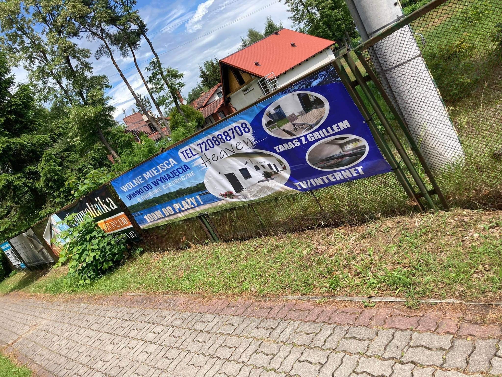 Baner reklamowy domków letniskowych 'Heaven' z informacjami o wynajmie, tarasie z grillem i dostępności TV/Internet, umieszczony na ogrodzeniu z siatki, widoczny z chodnika.