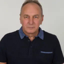 Portret mężczyzny o niebieskich oczach w ciemnej koszulce polo z jasnym kołnierzykiem, na jednolitym tle. Ujęcie centralne, neutralny wyraz twarzy.