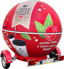 Mobilna kawiarnia w kształcie kuli na przyczepie, w kolorze czerwonym z białymi napisami 'Coffee & more' oraz grafiką filiżanki kawy, z widocznymi kołami i tablicą rejestracyjną.