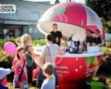 Mobilna kawiarnia w kształcie owocu, obsługiwana przez osobę wewnątrz, otoczona klientami z balonami na zewnątrz, ustawiona na trawniku przed budynkiem.