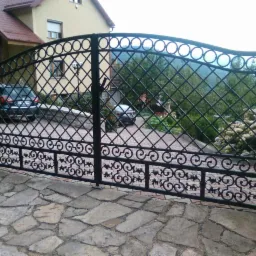 Czarna, dwuskrzydłowa brama z kutymi zdobieniami, widoczna z zewnątrz posesji. W tle dom z czerwonym dachem i podjazd wyłożony kostką.