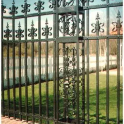 Czarne, kute ogrodzenie z dekoracyjnymi elementami i złotymi zdobieniami na tle zielonego trawnika i błękitnego nieba. Ogrodzenie osadzone na ceglanym murku.