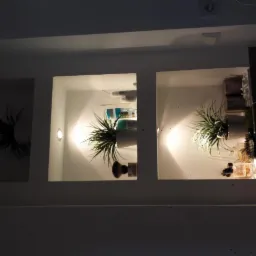 Podświetlane wnęki w ścianie, w dwóch z nich rośliny w doniczkach, w jednej kosmetyki. Oświetlenie punktowe tworzy ciepły klimat.
