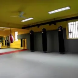 Wnętrze sali treningowej sztuk walki z żółtymi ścianami, rzędem czarnych worków bokserskich pod oknami z kratami i pojedynczym czerwono-białym workiem wiszącym w rogu.