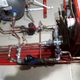 Złożona instalacja hydrauliczna z miedzianymi rurami, pompami obiegowymi (jedna z niebieską obudową), zaworami i zbiornikiem wyrównawczym, zamontowana na ścianie z poziomnicą w tle.