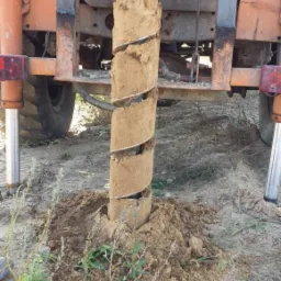 Wiertło do studni z wydobytą ziemią w kształcie spirali, osadzone w pomarańczowej maszynie wiertniczej na zewnątrz, z ziemią i roślinnością w tle.