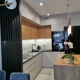 Nowoczesna kuchnia w kształcie litery L z drewnianymi górnymi szafkami, białymi dolnymi szafkami, blatem z efektem marmuru i czarną armaturą, oświetlona wiszącymi lampami nad wyspą kuchenną,...