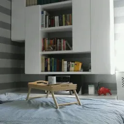 Białe szafki wiszące nad łóżkiem z książkami, drewniana taca śniadaniowa z kubkiem na łóżku w pokoju z szaro-białymi pasami na ścianie.