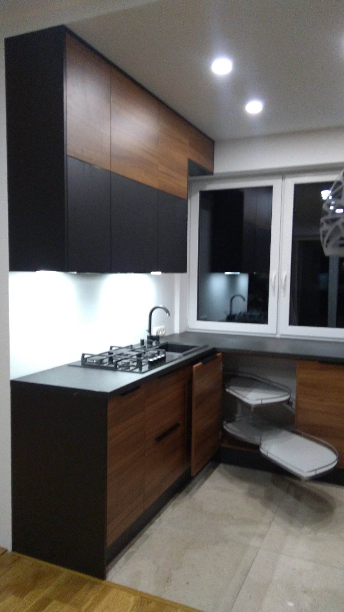 Nowoczesna zabudowa kuchenna z ciemnymi szafkami dolnymi i drewnopodobnymi górnymi, oświetlenie LED pod szafkami, narożny system półek obrotowych.