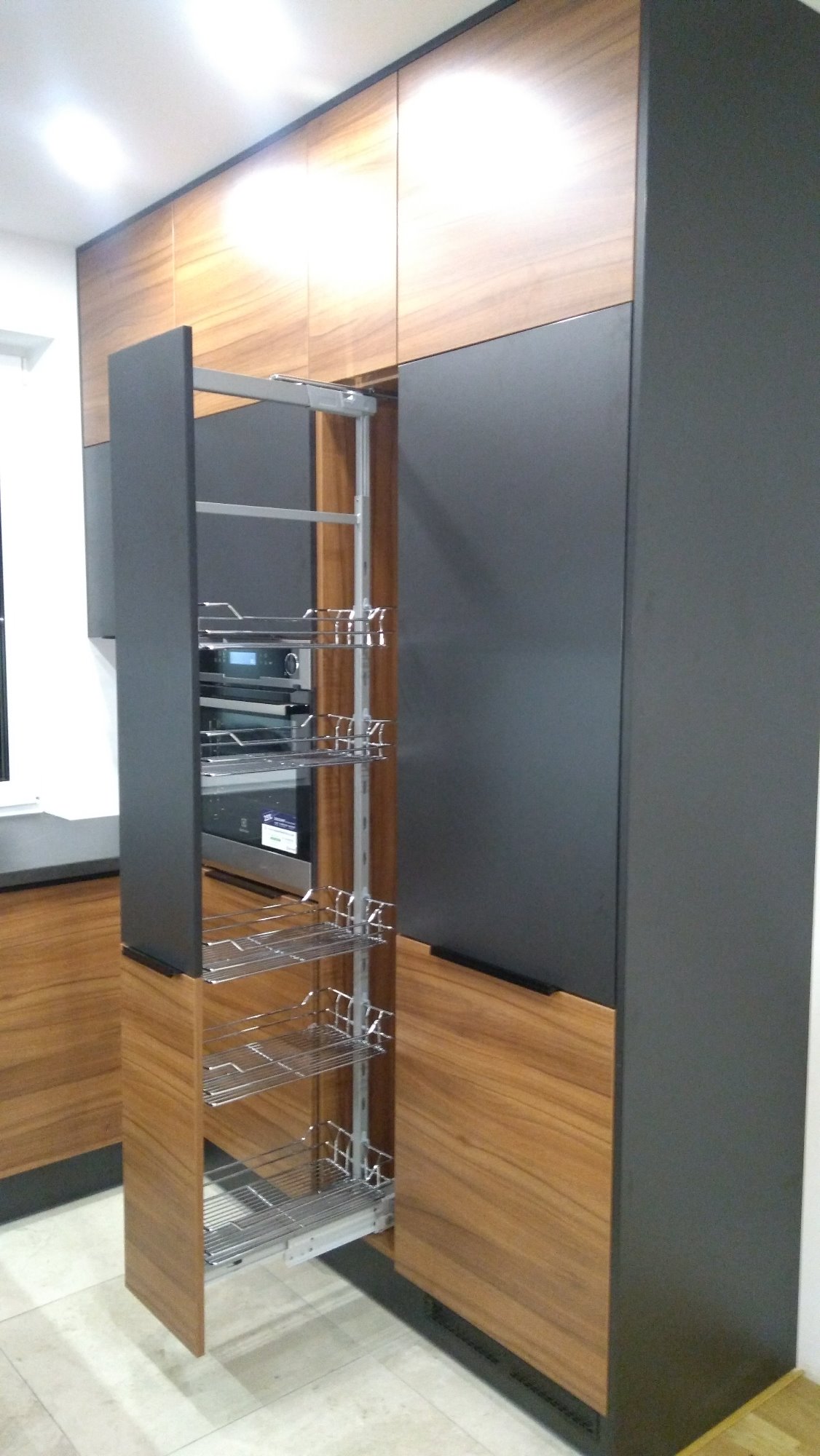 Wysoka szafka kuchenna z wysuwanym, metalowym systemem półek cargo, widoczny piekarnik i mikrofalówka w zabudowie, fronty w kolorze drewna i ciemnoszarym macie.