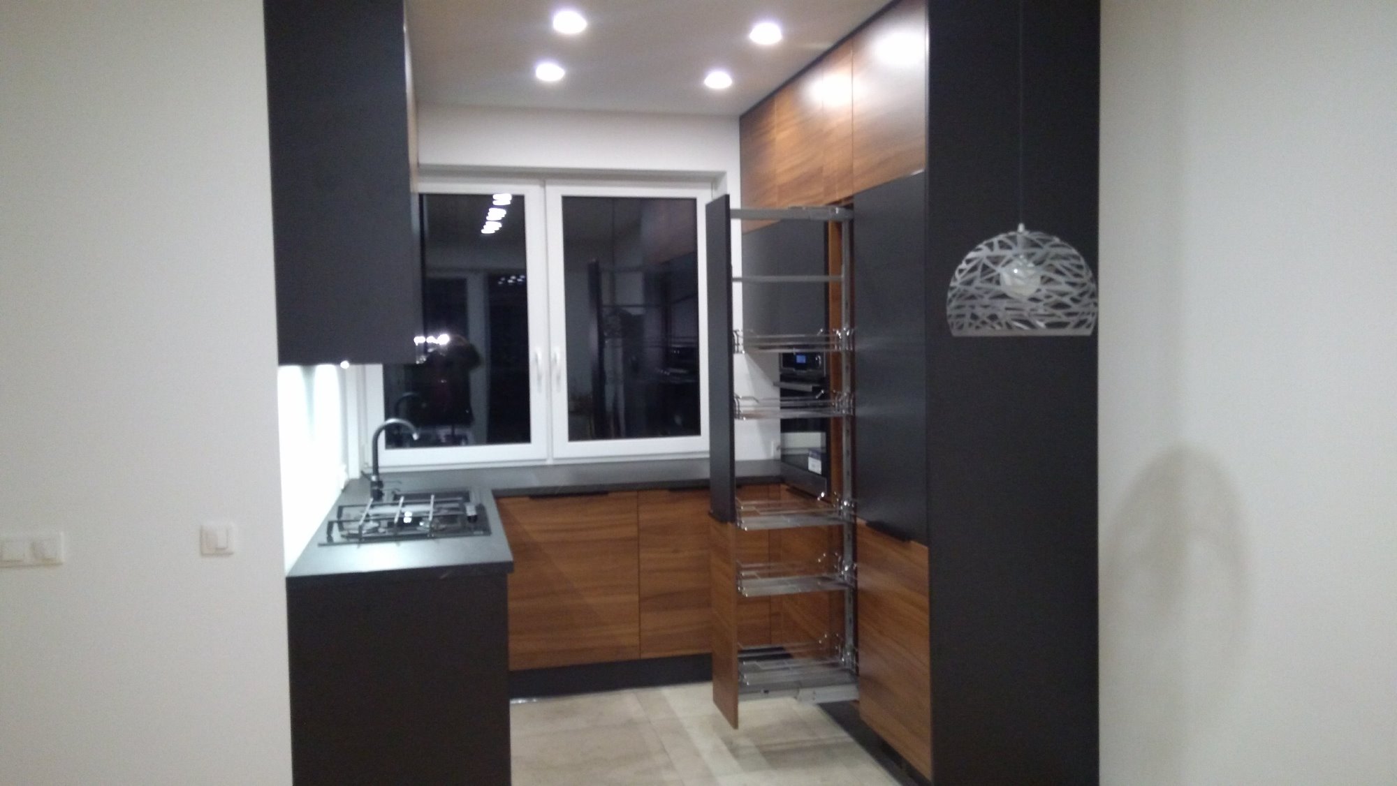 Nowoczesna kuchnia z otwartą szafką cargo, ciemnymi frontami i drewnianymi akcentami, oświetlona punktowymi lampami sufitowymi.