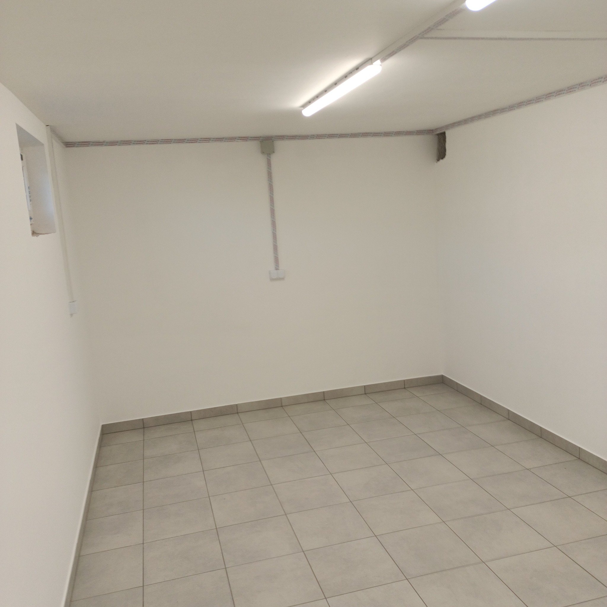 Puste, świeżo wyremontowane pomieszczenie z szarymi płytkami podłogowymi, białymi ścianami i widoczną instalacją elektryczną pod sufitem.
