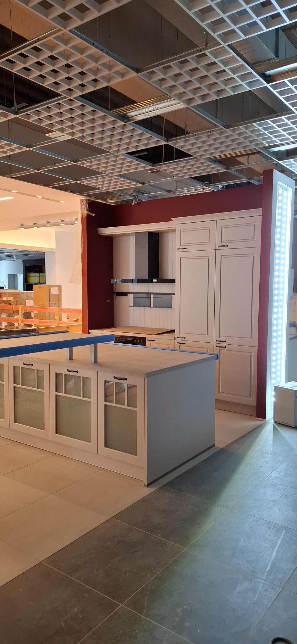 Wnętrze w trakcie prac wykończeniowych: zamontowane białe szafki kuchenne z wyspą na tle bordowej ściany, widoczna surowa instalacja sufitowa.